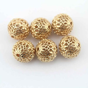 Puede incluir: Seis cuentas de metal de color dorado con un diseño decorativo, intrincado y calado. Las cuentas son redondas y tienen un agujero en el centro para enhebrar.