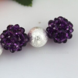 May include: Close-up of beads in a row. Two dark purple, faceted beads are on either side of a silver, textured bead. The purple beads have a clustered, floral-like shape. The silver bead has a speckled appearance.