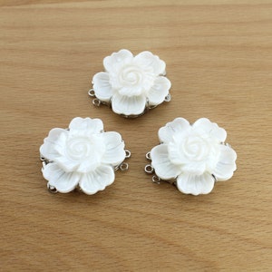 Puede incluir: Tres adornos blancos en forma de flor con un diseño detallado de pétalos y conectores de metal plateado. Las flores están dispuestas sobre una superficie de madera marrón claro, mostrando sus delicadas características.