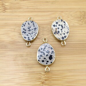 May include: Three oval-shaped dalmatian jasper gemstone pendants with gold-toned metal connector loops. The gemstones have a white base with black spots.