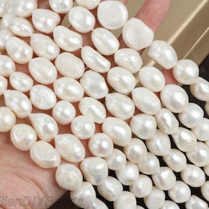 May include: A strand of white baroque pearls, with a slightly irregular shape, arranged in a row on a hand. The pearls are smooth and lustrous.