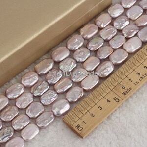 May include: A strand of light purple baroque freshwater pearls, measuring approximately 1.5 cm in length, arranged in a row on a wooden ruler. The pearls have an iridescent sheen and are oval-shaped.