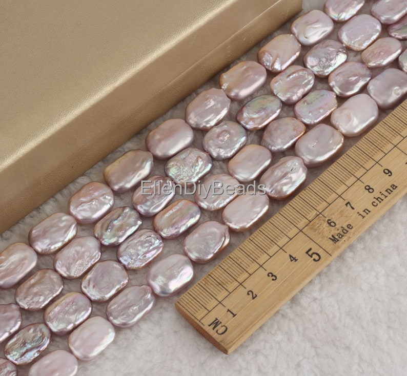 May include: A strand of light purple baroque freshwater pearls, measuring approximately 1.5 cm in length, arranged in a row on a wooden ruler. The pearls have an iridescent sheen and are oval-shaped.