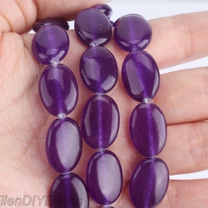 Puede incluir: Una hebra de cuentas de amatista de forma ovalada en un color morado intenso. Las cuentas son lisas y pulidas, con un ligero brillo.