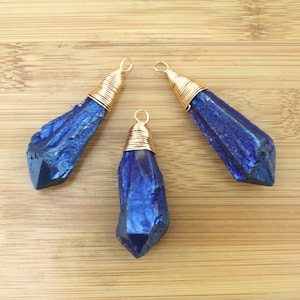 May include: Three blue crystal pendants with gold wire wrapping. The crystals have a rough, natural texture and a deep blue colour with iridescent highlights. Each pendant has a gold-coloured loop at the top for hanging.