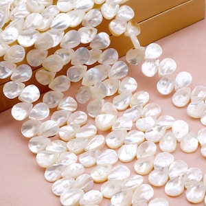 May include: Close-up of a strand of pearly white, teardrop-shaped beads. The beads are strung together, creating a flowing, organic pattern. The beads are arranged on a light pink surface, with a gold-colored box in the background.