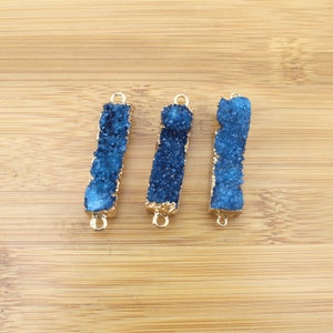 Puede incluir: Tres colgantes de barra de piedra de ágata azul con bucles de metal dorado. Las piedras son de color azul intenso y tienen una superficie rugosa y texturizada.