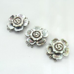 May include: Three flower-shaped jewelry clasps. Each clasp features a mother-of-pearl design with iridescent hues of gray, green, and purple. The clasps have a silver-colored metal base with multiple loops for attachment.