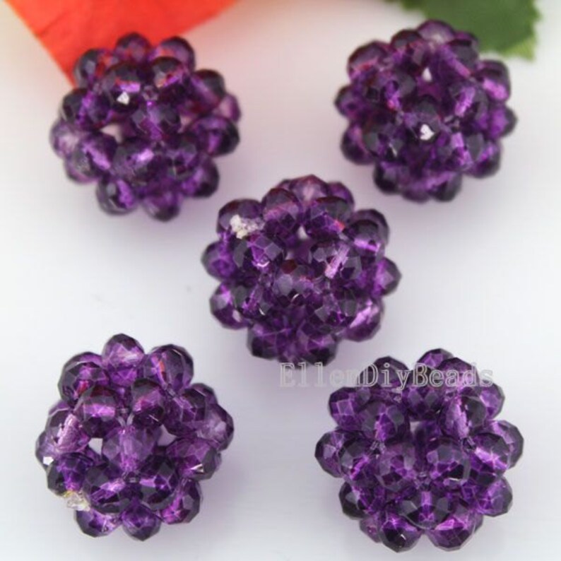 May include: Five faceted, spherical beads in a deep purple hue. Each bead is composed of numerous small, geometric facets, creating a sparkling, textured surface. The beads are clustered together, resembling small, decorative balls.