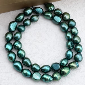 May include: A double strand necklace of large, irregularly shaped, dark teal pearls. The pearls have an iridescent sheen, reflecting green and blue. The necklace is displayed on a white surface.