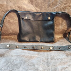 May include: Black leather fanny pack with a flap closure secured by a single snap. The adjustable strap is gray and adorned with silver-toned rivets. The bag is designed to be worn around the waist or across the body.