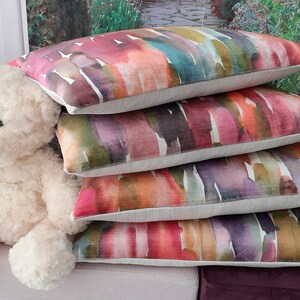 May include: A stack of four rectangular throw pillows featuring a multicolored watercolor-style print with shades of pink, orange, teal, purple, and green on a light beige fabric base, positioned next to a cream-colored plush teddy bear.