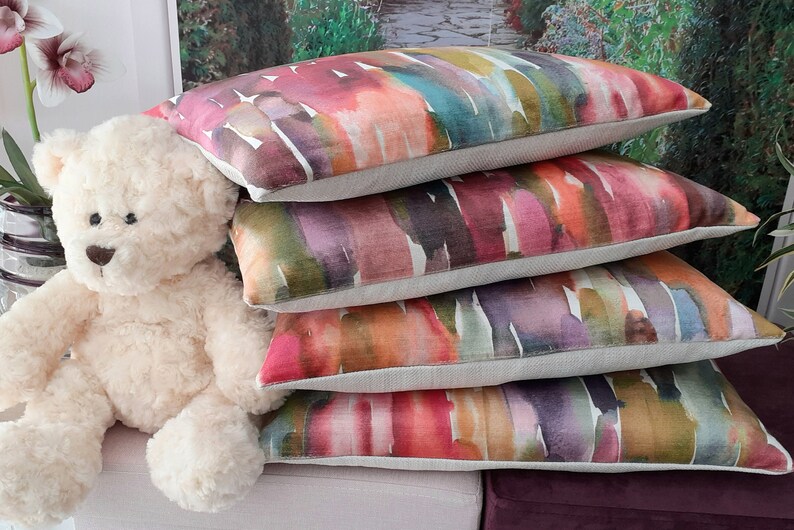 May include: A stack of four rectangular throw pillows featuring a multicolored watercolor-style print with shades of pink, orange, teal, purple, and green on a light beige fabric base, positioned next to a cream-colored plush teddy bear.