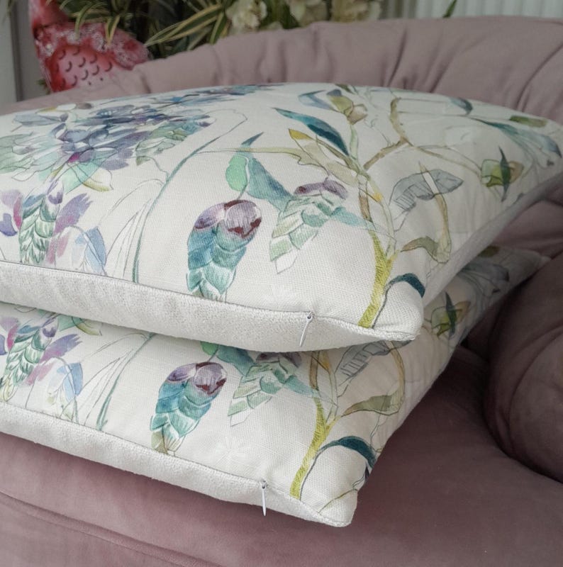 May include: Two white cushion covers with a floral pattern featuring green, purple, and yellow watercolour-style flowers. The cushions are stacked on top of each other on a pink sofa.