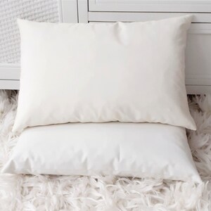 May include: Two rectangular pillows with smooth, solid off-white fabric covers. The pillows are stacked one on top of the other, featuring a clean, plain design with no visible patterns, textures, or embellishments on the surface of the fabric.