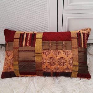 May include: A rectangular throw pillow with a patchwork design featuring various shades of brown, red, and gold. The pillow has a geometric pattern with circles and stripes.