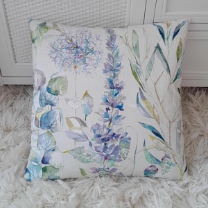 May include: A white cushion with a floral pattern featuring blue, green, and purple watercolour flowers. The cushion is made of a soft, textured fabric.