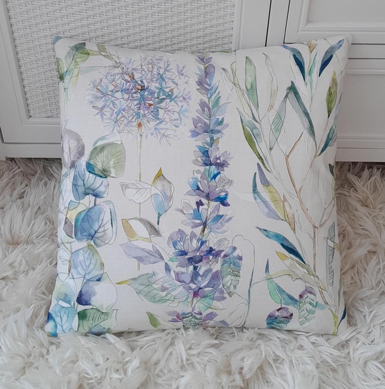May include: A white cushion with a floral pattern featuring blue, green, and purple watercolour flowers. The cushion is made of a soft, textured fabric.