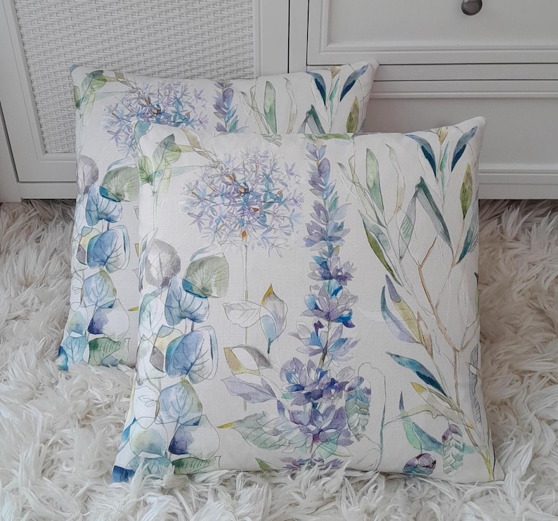 May include: Two decorative throw pillows with a floral pattern featuring watercolour-style blue, green, and purple flowers on a white background.