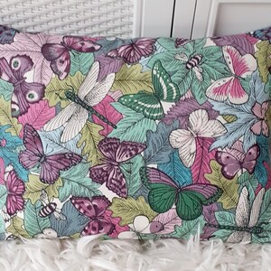 May include: A decorative cushion featuring a colourful butterfly and dragonfly design on a white background. The cushion is made of fabric and has a rectangular shape.