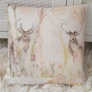 May include: A square throw cushion featuring a watercolour-style print of two deer standing among birch trees and a small rabbit in the grass. The design uses soft, muted tones of brown, green, and beige on a light, textured fabric background.