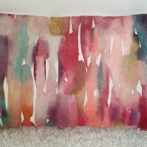 May include: A rectangular throw pillow featuring a vibrant watercolor print with vertical streaks of pink, magenta, deep red, teal, orange, and muted gold against a white background. The fabric has a visible woven texture throughout the design.