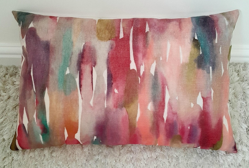 May include: A rectangular throw pillow featuring a vibrant watercolor print with vertical streaks of pink, magenta, deep red, teal, orange, and muted gold against a white background. The fabric has a visible woven texture throughout the design.