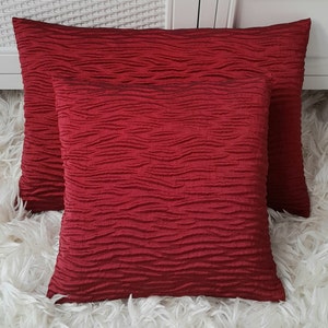 May include: Two red decorative throw cushions with a textured, wavy pattern. The cushions are arranged on a white fluffy surface.