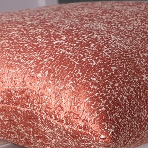 May include: A close-up of a copper-coloured throw pillow with a white and copper patterned design. The pillow is made of a soft, textured fabric.