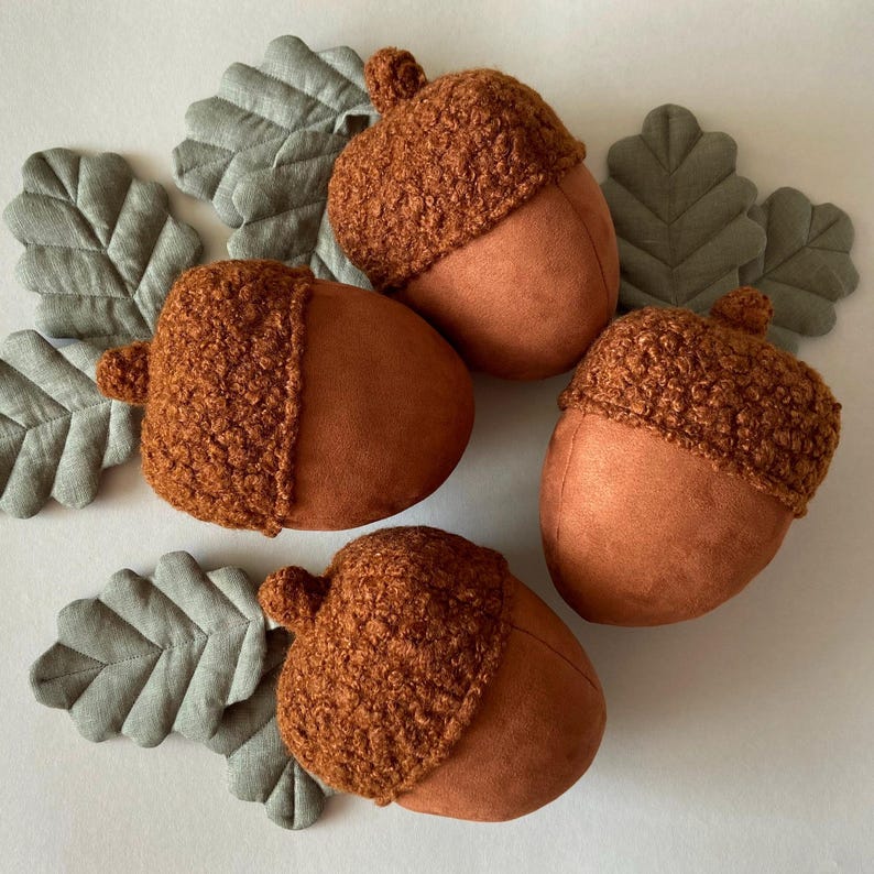 May include: Four brown felt acorns with fuzzy brown tops. The acorns are arranged on a white surface with green felt leaves.