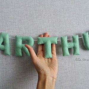 May include: A green felt banner with the name "ARTHUR" spelled out in individual letters. The letters are strung together with white thread.