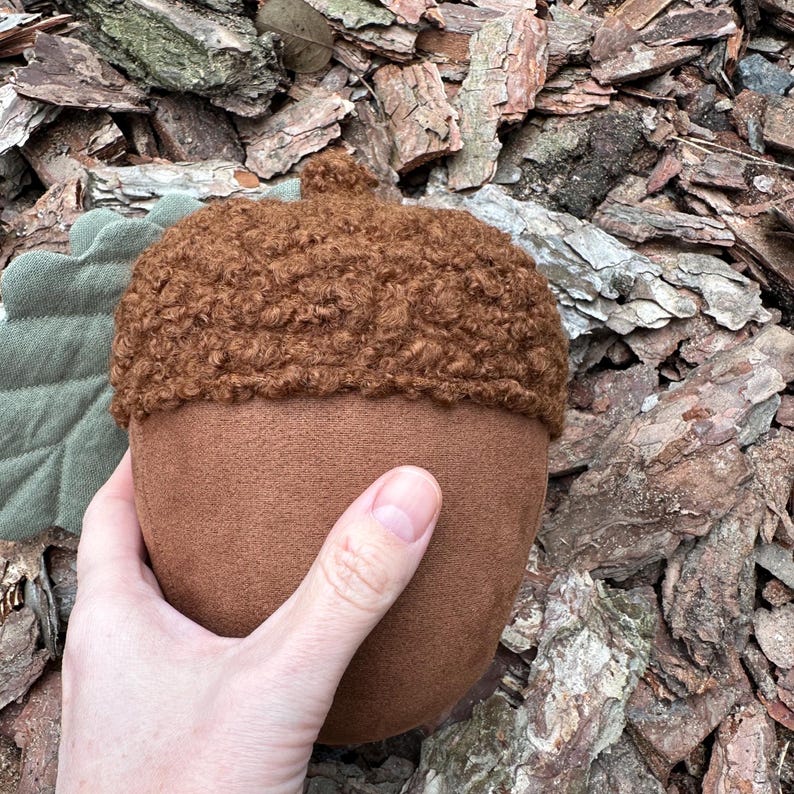 May include: A brown plush acorn with a fuzzy brown cap. The acorn is held in a hand against a background of brown wood chips.