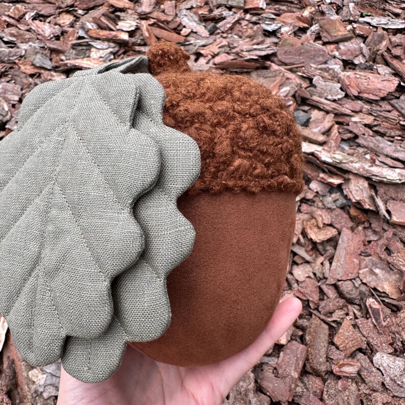 May include: A plush acorn toy with a brown felt body and a fuzzy brown cap. The acorn is attached to a green felt leaf.