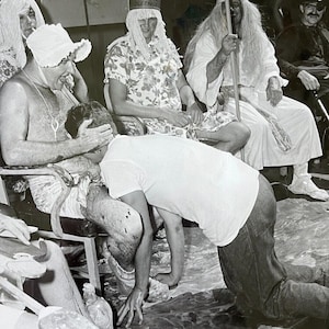 May include: A group of people dressed in costumes, including a person wearing a white crown with the word "Queen" on it, a person wearing a white robe and a long white beard, and a person wearing a white shirt and jeans. The people are sitting on chairs and appear to be having fun.