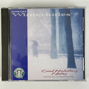 May include: A Starbucks "Winterludes" CD in a clear plastic case. The cover features a winter scene with a silhouetted figure and trees. Text includes "Cool Holiday Notes" and "Seasonal Jazz and Popular Favorites."