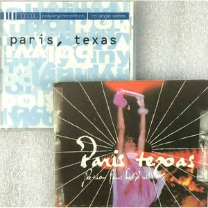 May include: A CD single by the band Paris Texas. The front cover features a black and white photo of a person holding a pink heart-shaped object. The text "Paris Texas" is written in white on a black background. The text "Action fans help us!" is written in white on a black background.