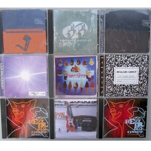 May include: A collection of nine compact discs in clear plastic cases. The discs are arranged in three rows of three. The discs feature various album covers with different designs and colors. The top row features a disc with a blue and orange cover, a disc with a green cover, and a disc with a black and white cover. The middle row features a disc with a purple cover, a disc with a yellow cover, and a disc with a black and white cover. The bottom row features a disc with a red cover, a disc with a blue cover, and a disc with a red cover.