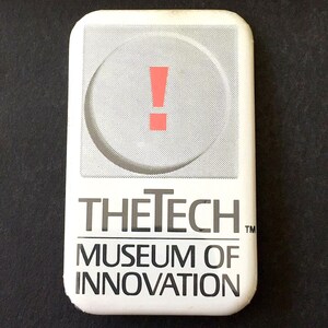 May include: A rectangular white button with rounded corners. The top section features a gray circle with a red exclamation point. Below, the text reads "THE TECH MUSEUM OF INNOVATION" in black.