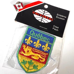 May include: A shield-shaped embroidered patch featuring the word Québec, three yellow fleurs-de-lis, a yellow lion, and green maple leaves on a blue, red, and yellow background, packaged with a small Canadian flag lapel pin.