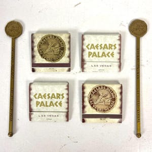 Könnte beinhalten: Eine Auswahl an Vintage-Erinnerungsstücken von Caesars Palace Las Vegas. Das Bild zeigt zwei goldfarbene Rührstäbchen und vier Streichholzschachteln. Die Streichholzschachteln tragen das Caesars Palace-Logo und den Text. Die Rührstäbchen haben ebenfalls das Caesars Palace-Logo.