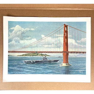 May include: A vintage print of the Golden Gate Bridge in San Francisco, California. The iconic red bridge spans the water with a naval aircraft carrier sailing beneath it. The sky is blue with fluffy white clouds.