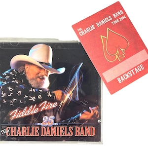 May include: A red backstage pass with the text "The Charlie Daniels Band Tour 2008" and a gold spade symbol. The pass has a blank white space for a name. A CD case with a picture of Charlie Daniels playing a fiddle is in the background.