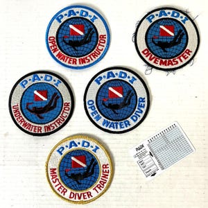 May include: A collection of embroidered PADI patches, featuring the PADI logo, a diver silhouette, and text indicating different diving certifications like "Open Water Instructor" and "Divemaster." A dive table is also included.