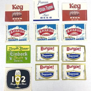 Peut inclure: Collection d'étiquettes de bière vintage avec différentes marques. Les étiquettes incluent "Keg", "Olde Tyme Lager", "Bavarian Weiss", "Einbock Draft" et "Burgie!". Les motifs comprennent des scènes de montagne, des tonneaux et une typographie classique. Les étiquettes sont rectangulaires et de couleurs variées.