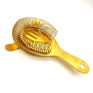Puede incluir: Un colador de cóctel de color dorado con un diseño de resorte en espiral alrededor de la cabeza del colador. El colador tiene un mango largo con un agujero en el extremo para colgarlo. La herramienta de bar está diseñada para colar hielo y otros ingredientes de las bebidas mixtas.