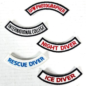 May include: A collection of embroidered patches with curved designs. The patches feature text such as "U/W PHOTOGRAPHER," "INTERNATIONAL COLLEGE," "NIGHT DIVER," "RESCUE DIVER," and "ICE DIVER." The patches have a white background with black and red text.