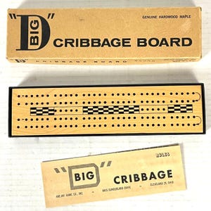May include: A cribbage board game set with a wooden board and box. The box reads "BIG CRIBBAGE BOARD" and "GENUINE HARDWOOD MAPLE". The board has black and white checkered patterns and numerous holes for pegs. A rules card is also included.
