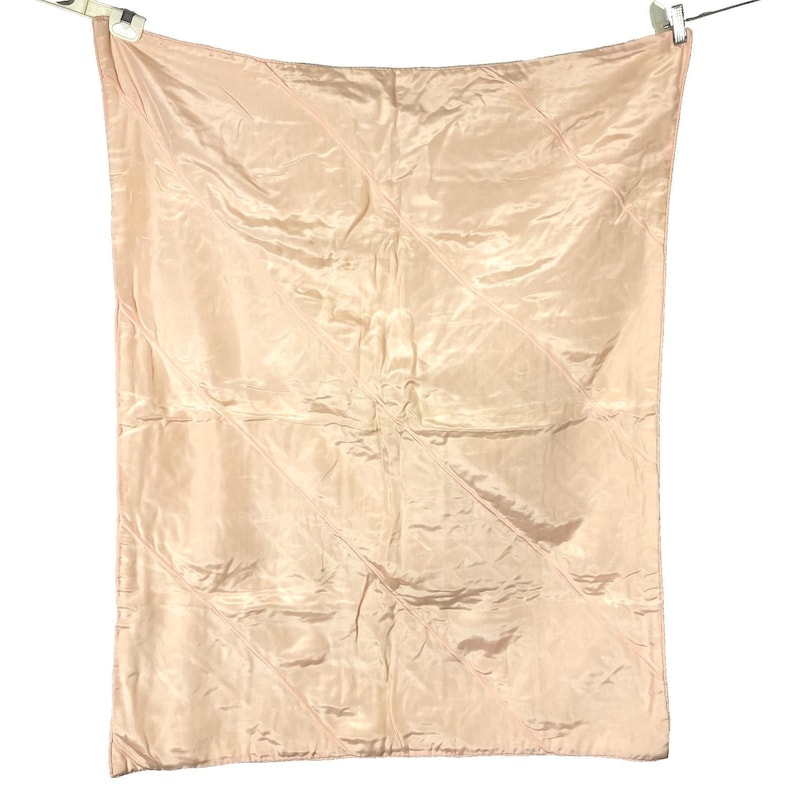 May include: A light pink, quilted blanket hanging from a clothesline. The blanket has a diamond pattern and a shiny, satin-like texture. The blanket is rectangular and appears to be made of a soft material.