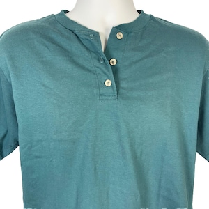 May include: Teal-colored Henley shirt with a classic collar and three beige buttons. The short-sleeved shirt is made of a soft, breathable fabric. The shirt is displayed on a mannequin.