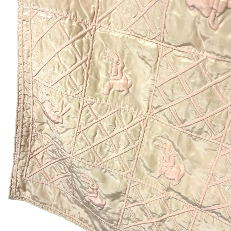 May include: A quilted blanket with a repeating pattern of pink rabbits on a cream-colored background. The blanket has a grid-like design with raised pink stitching. The rabbits are in various poses, adding a whimsical touch to the textile.